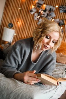 Woman relaxing and reading book on cozy bed - Wooden wall and photos with lights - blurred background - vertical photo