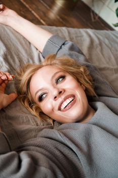 Beautiful young woman in gray sweater laughing lying in bed in her cozy bedroom. Cute girl relaxing at home. Happy morning