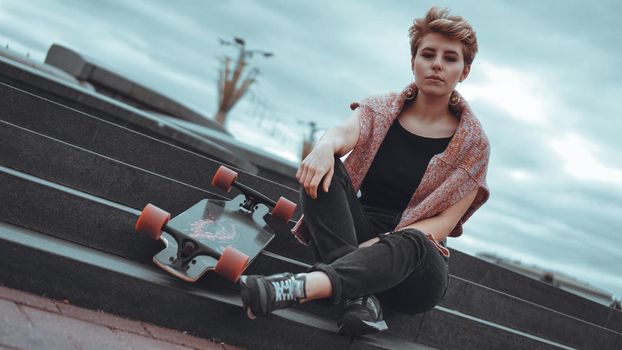 A beautiful young girl holding a skateboard while sitting in skatepark at the city