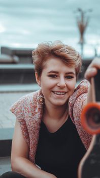 A beautiful young girl holding a skateboard while sitting in skatepark at the city