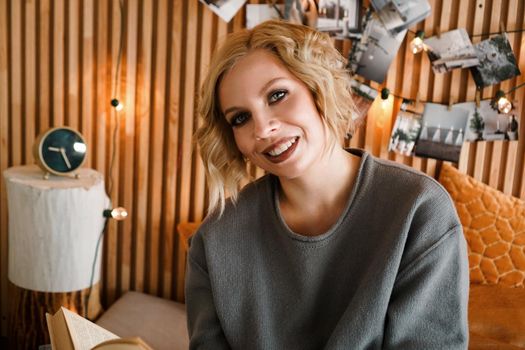 Happy Woman looking to the camera, relaxing and reading book on cozy bed - Wooden wall and photos with lights - blurred background