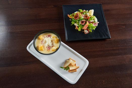 What to choose Bowl of cheese cream soup with croutons served with a plate of prosciutto salad top view