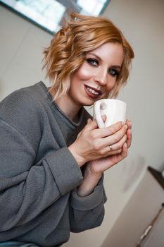 Smiling blonde drinking cappuccino, holding coffee cup. Vertical photo. Stay at home, cozy kitchen