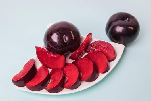 On a white saucer there is a large plum cut into wedges, next to it there are two more plums. The pulp of red berries. Close-up.
