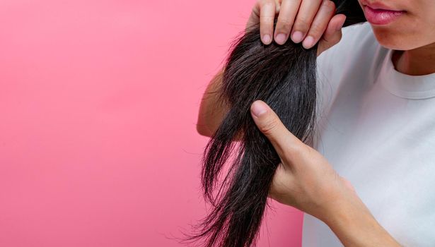 Asian woman with dry lips holding damaged hair on pink background. Dry and brittle black long hair needs shampoo and conditioner for spa treatment. Hair loss and thin hair problems in women. 