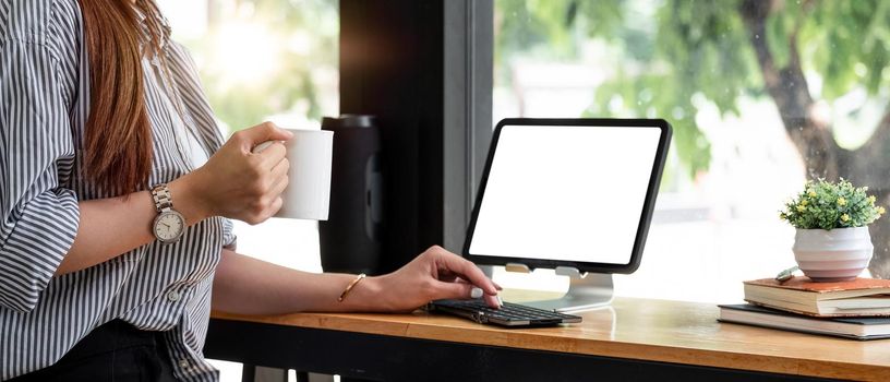 Woman hand holding cup of coffee and using digital tablet. Female working at coffee shop freelance concept.