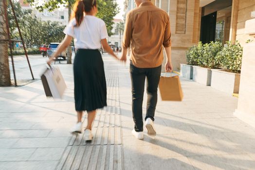 Lower body section of a young tourist couple walking by store windows and holding paper shopping bags in a destination city.