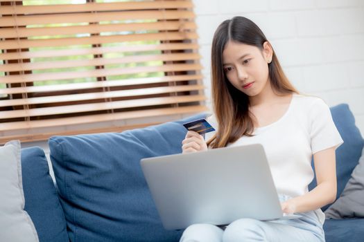 Young asian woman sit on sofa using laptop computer shopping online with credit card buying to internet, happy girl payment with e-business on couch, purchase and payment, business concept.