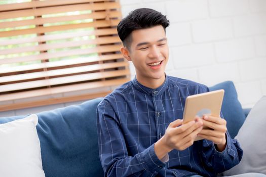 Young attractive asian man resting using browsing tablet computer on sofa at home, happy male sitting on couch relax reading digital gadget at house, communication and lifestyle concept.