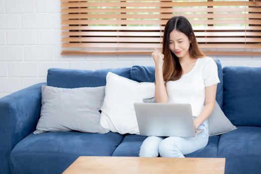 Young asian business woman smile and work from home with laptop computer online to internet on sofa in living room, freelance girl using notebook on couch with comfort, new normal, lifestyle concept.