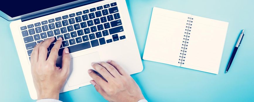 Hand of businessman working on laptop computer with notebook and cup of coffee on desk in office, hand of person typing keyboard with book and pen, top view, flat lay, business concept.