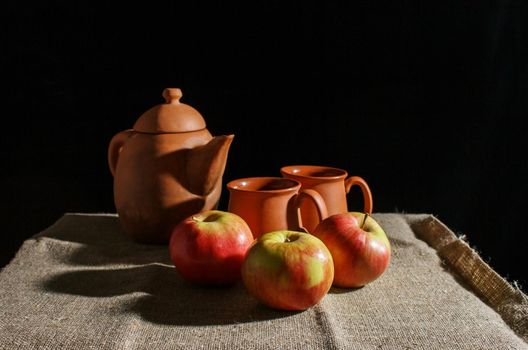 still life with a clay teapot, two cups and three red apples. indoor closeup on black background