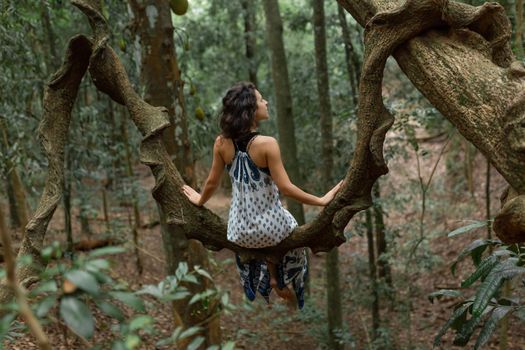 The girl sits on a huge liana tree branch in the jungle.