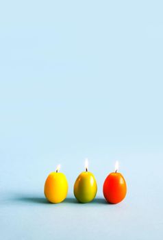 Traditional Easter decor. Bright burning paraffin candles in the shape of colorful eggs on blue background.