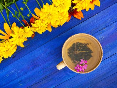 Natural herbal tea with purple fresh flowers and leaves of medical fireweed plant n ceramic cup on blue wooden boards outdoors.