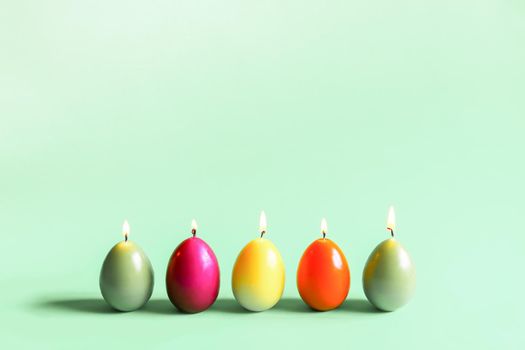 Traditional Easter decor. Group of bright burning paraffin candles in the shape of colorful eggs on soft green background.