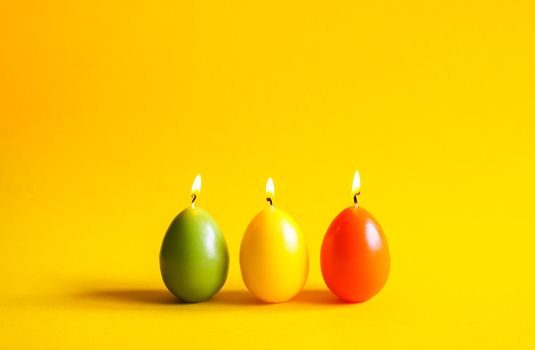 Traditional Easter decor. Group of bright burning paraffin candles in the shape of colorful eggs on soft warm yellow background.