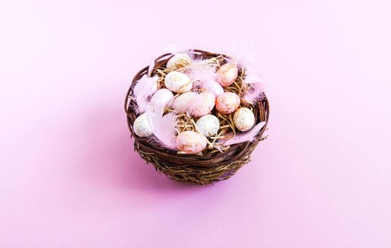 Easter composition with traditional decor. Small decorative colorful eggs and soft feathers in a wicker basket on pink light background.