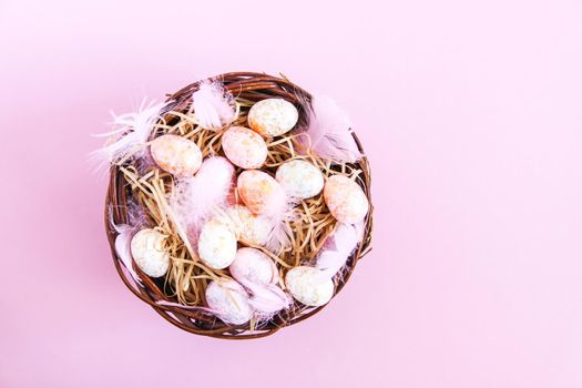 Easter composition with traditional decor. Small decorative colorful eggs and soft feathers in a wicker basket on pink light background.