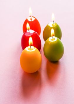 Traditional Easter decor. Group of bright burning paraffin candles in the shape of colorful eggs on soft pink background.