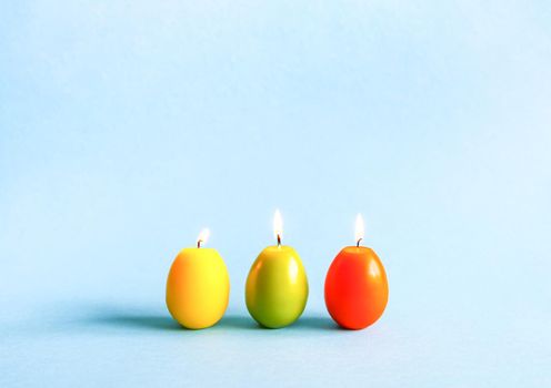 Traditional Easter decor. Bright burning paraffin candles in the shape of colorful eggs on blue background.