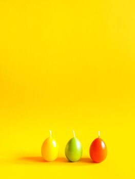 Traditional Easter decor. Group of bright burning paraffin candles in the shape of colorful eggs on soft warm yellow background.