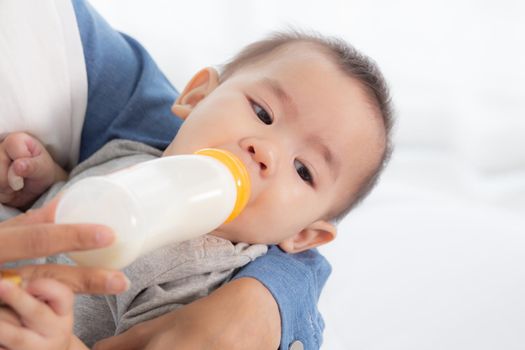 Young asian mother embracing and feeding little baby girl with bottle of milk at home, newborn innocence drinking with mom satisfied, relationship and bonding of mum and child, family concept.