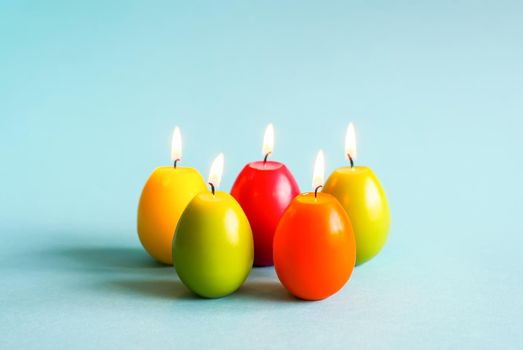 Traditional Easter decor. Bright burning paraffin candles in the shape of colorful eggs on blue background.