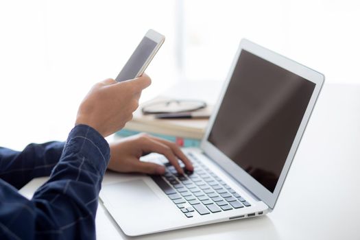 Hand of young man working with laptop computer and smartphone mockup on desk at home, notebook and phone display blank screen, freelance look message to internet, business and communication concept.