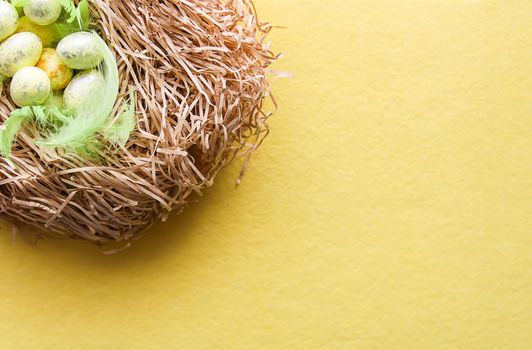 Easter composition with traditional decor. Small decorative colorful eggs on soft artificial hay on soft yellow background.