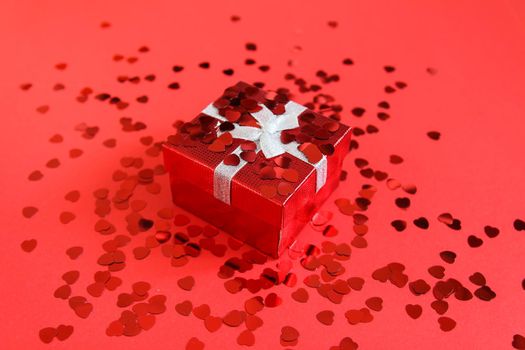 Small red gift box with silver ribbon on bright background with decorative small heart tinsel.