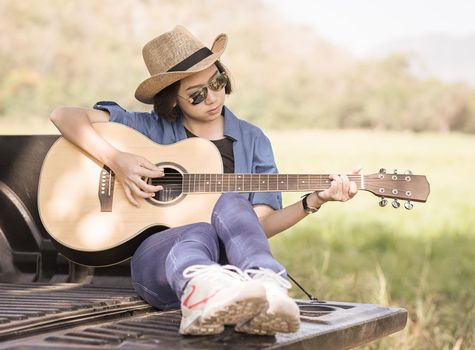 Young asian women short hair wear hat and sunglasses playing guitar ,sit on pickup truck in countryside Thailand