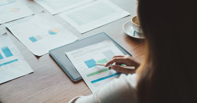 Close up Businesswoman hand holding pen and pointing at financial paperwork and using digital tablet.