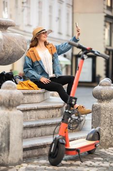 Young woman, sitting on staircase of old historical building in city center, taking picture with her phone. Female traveler exploring Ljubljana's old town on electric scooter. Summer leisure