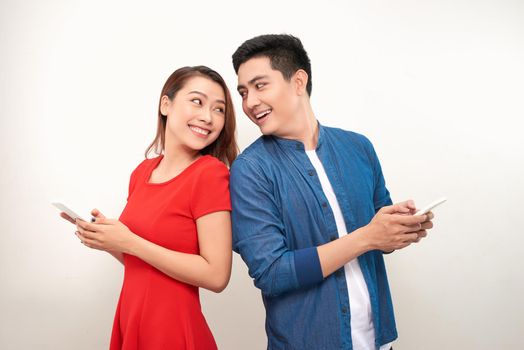 Beautiful young asian couple in casual clothes is using smartphones, looking at camera and smiling, standing back to back against white background