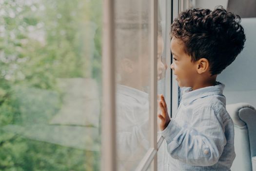 Side view of lovely afro american kid toddler waiting for mom, dressed in blue shirt, looking out of window through his reflection leaning on glass with hands and smiling. Children leisure time indoor