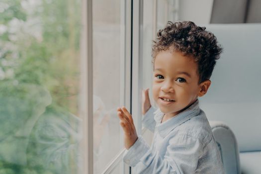 Portrait of mixed race curly haired baby boy in light blue colored shirt smiling at camera while leaning on window glass and playing alone at home, child toddler spending time indoors without parents