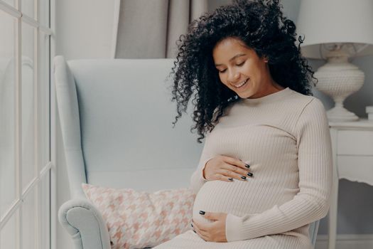 Beautiful mixed race pregnant woman in casual dress sitting in armchair by window tenderly embracing belly thinking about new baby to come. Happy maternity and pregnancy concept