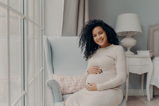Happy young pregnant afro american lady in dress preparing soon to be mother, sitting on edge of cozy armchair in front of big spacious window and smiling at camera. Pregnancy and maternity concept