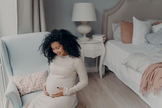Lovely young future mother in white dress with curly hair, looking down at her belly and smiling, trying to come up with name for baby and planning out nursery room. Happy pregnancy concept