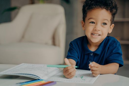 Cute mixed race small boy in polo tshirt painting coloring book with felt tip pens, sitting by table in living room against blurred background with armchair. Children leisure time at home concept