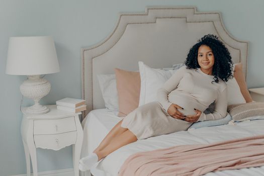 Happy lovely pregnant beautiful afro american woman in casual white dress with curly hair resting on bed in light interior bedroom at home, touching her belly and smiling at camera. Pregnancy concept