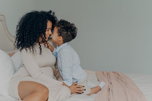 Tender moment of young family, afro american pregnant mother and son sitting on bed,touching each other with noses, white wall background in bedroom interior. Happy parenting and motherhood concept