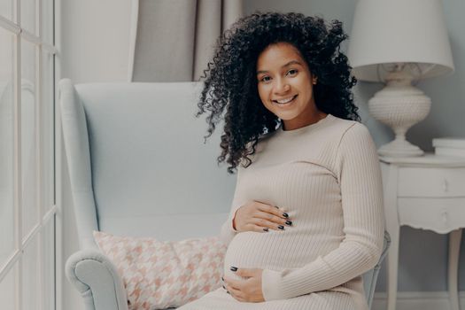 Preparing for motherhood. Beautiful pregnant african woman with curly hair sitting on armchair in light room at home and holding her belly, expecting baby and proud to be future mother