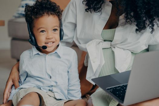 Happy afro american child with curly hair looking aside and smiling, wearing headset while having online meeting with dad, using laptop with his mother, boy talking with family through webcam