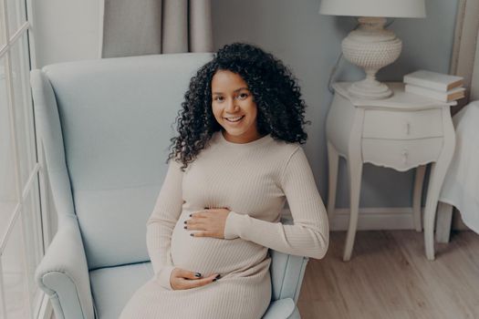 Portrait of happy smiling elegant woman future mom in light colored dress sitting sideways on armchair tenderly touching tummy next to white nightstand with lamp. Enjoying pregnancy time concept