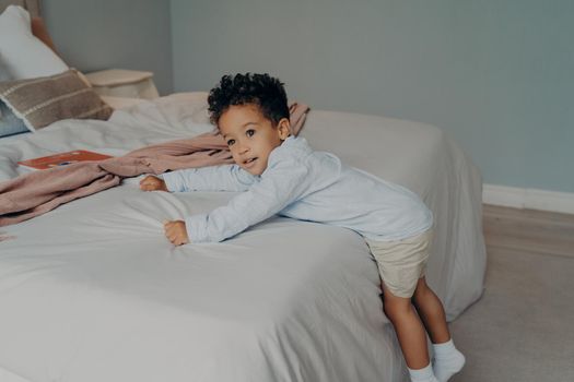 Sweet little afro american kid boy in stylish cotton clothes trying to climb on big white soft bed, holding onto bedsheet while playing in bright bedroom at home, preschool child spending leisure time