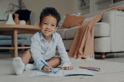 Little cutie curly haired afro baby boy sitting on floor of modern living room interior next to sofa and drawing with colorful felt tip pens in album. Creative hobby for kids and playtime activity