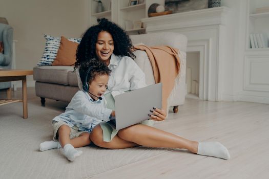 Young positive mixed race mother and cute boy son having video call with relatives, father or grandparents, sitting with laptop on floor and smiling, child wearing headset and pointing at screen