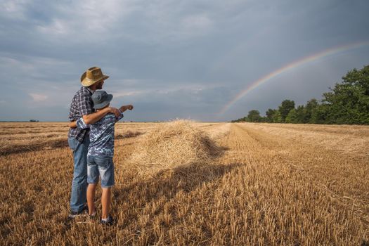 Father and son are standing in their wheat field after successful harvest. They are looking at rainbow in the sky.